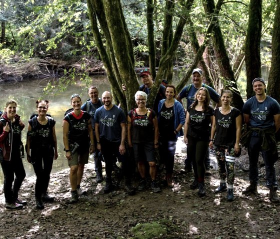 12 Personen in Wanderkleidung stehen neben einem Fluss im Wald und schauen lachend in die Kamera nach oben.
