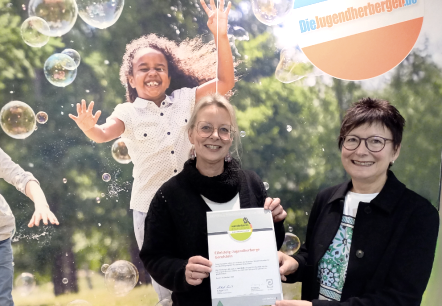 The photo shows two women proudly holding a certificate in front of a background depicting a young, cheerful person surrounded by soap bubbles.The scene shows two women posing for a photo, one of them holding the certificate they are presenting. The background shows a large-format image of a child smiling happily at the camera surrounded by soap bubbles. The background is a green, nature-like backdrop. The women stand in front of the background, with the woman on the right holding the certificate for the camera, while the woman on the left is also involved. The alignment of the photo is good and the main focus is on the women and the certificate. The background serves as a complementary backdrop.The two main characters are two middle-aged women. The woman on the left has blonde hair, wears glasses, and a black sweater. She holds the certificate with a smile. The woman on the right has short brown hair, glasses, and a dark blazer. She is also smiling. The background shows a happy child playing in soap bubbles., &copy; Touristik GmbH Gerolsteiner Land