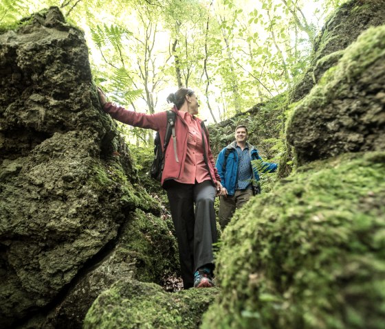 Twee mensen lopen over een met mos begroeid, rotsachtig pad in het bos. Ze dragen outdoorkleding en genieten van de natuur., &copy; Eifel Tourismus GmbH, Dominik Ketz