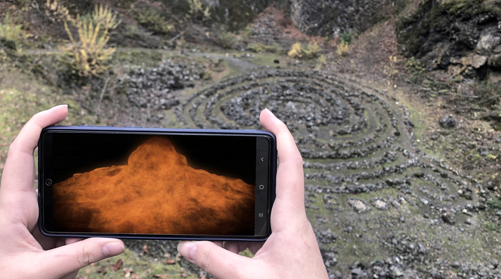 Two hands hold a smartphone across the abyss of a volcano. The volcanic explosion is visualised on the screen.