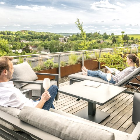 Ein Mann und eine Frauen sitzen entspannt auf der modern möblierten Dachterasse eines Hotels, trinken Wein und schauen in die grüne Landschaft.