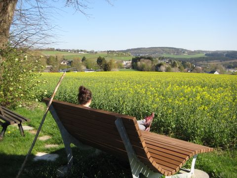 Person sitzt auf einer Bank mit Blick auf ein Rapsfeld und eine hügelige Landschaft.