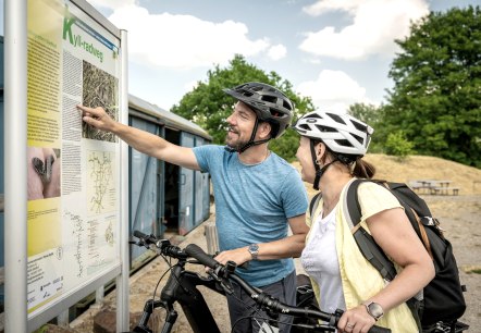Zwei Radfahrer mit Helmen studieren eine Infotafel am Kyll-Radweg. Sie l&auml;cheln und zeigen auf die Karte. Im Hintergrund sind B&auml;ume und ein blauer Container., &copy; Eifel Tourismus GmbH, Dominik Ketz