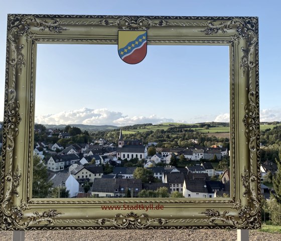 Golden picture frame with town coat of arms, framing the town of Stadtkyll and green landscape in the background. Clear sky and sunny weather., © Touristik GmbH Gerolsteiner Land, Leonie Post