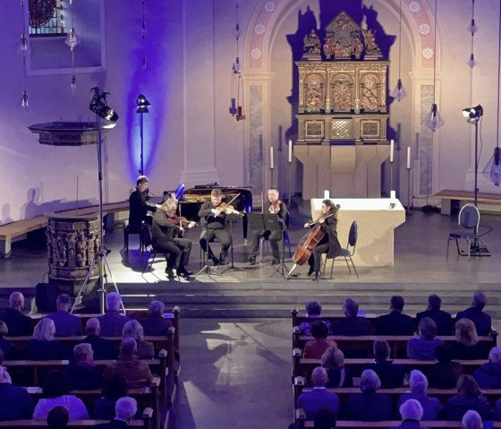 F&uuml;nf Musiker an Streichinstrumenten und Klavier musizieren vor dem Kirchenaltar. Davor die Sitzb&auml;nke voller Besucher.