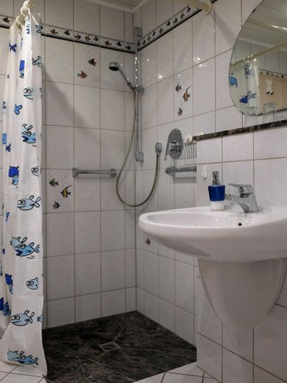 Ein Badezimmer mit einer Dusche und einem Waschbecken. Die Wände sind gefliest und mit einer bunten Duschvorhang mit Fischen dekoriert.