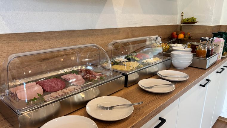 Ein Frühstücksbuffet mit verschiedenen Aufschnittsorten und Käse in Glaskästen. Neben dem Buffet stehen Teller und Besteck bereit.