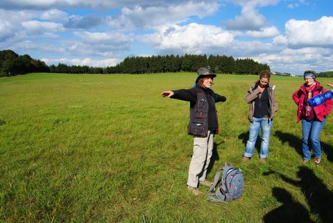 Drei Personen auf einer grünen Wiese, eine breitet die Arme aus. Sonniger Tag mit blauem Himmel und Wolken.