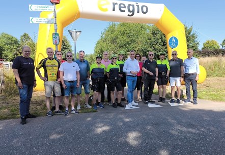 Talrijke personen, waarvan sommigen sportieve fietskleding dragen, staan op het fietspad onder een grote luchtboog van sponsor e-regio.