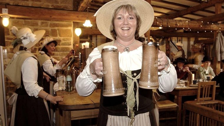 Frau in traditioneller Kleidung serviert Bierkrüge in einem rustikalen Restaurant mit Holzmöbeln und anderen Gästen im Hintergrund.