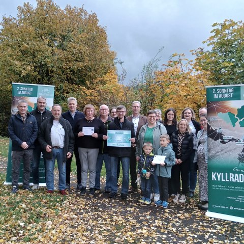 18 Personen stehen vor herbstlichen laubb&auml;umen auf einer schmalen asphaltierten Stra&szlig;e zusammen zwischen zwei Bannern f&uuml;r den Radaktionstag "Grenzenlos Kyllradweg" und l&auml;cheln in die Kamera. Drei Personen halten stolz jeweils einen Gewinn in den H&auml;nden.