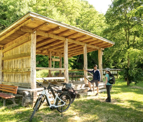 Zwei Radfahrer mit Helmen stehen neben einem h&ouml;lzernen Unterstand im Gr&uuml;nen. Fahrr&auml;der sind abgestellt, umgeben von B&auml;umen und Wiese., &copy; Eifel Tourismus GmbH, Dominik Ketz