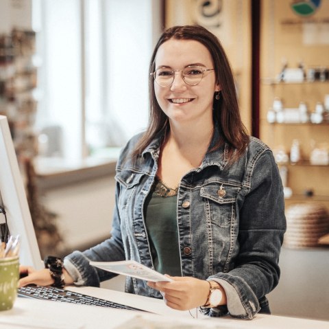 A woman stands at a high desk with a computer and keyboard in front of her and smiles at the camera. Behind her is the souvenir shop and some brochure racks from the tourist information office.