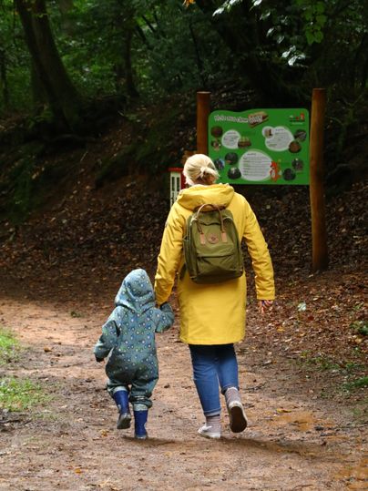 Eine Mutter mit ihrem Kleinkind an der Hand wandern durch einen Wald. Beide tragen Regenjacken.