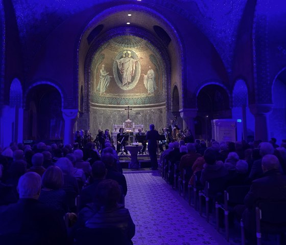 Schwach beleuchteter Innenraum der Erlöserkirche Gerolstein mit goldener Mosiakverzierung. Im Altarraum Musikanten und ein großes Publikum in den Sitzbänken.