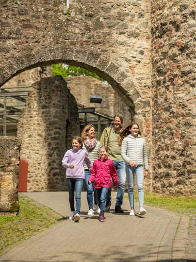 Eine fünfköpfige Familie geht unter einem Torbogen einer Burg auf einem gepflasterten Weg entlang.