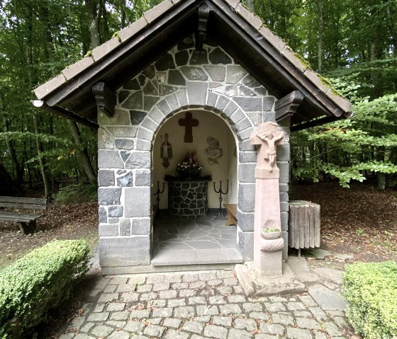 Die Arnulphuskapelle Walsdorf ist eine kleine Steinkapelle im Wald mit offenem Eingang und einem Kreuz im Inneren. Gepflasterter Weg f&uuml;hrt zur Kapelle., &copy; Touristik GmbH Gerolsteiner Land