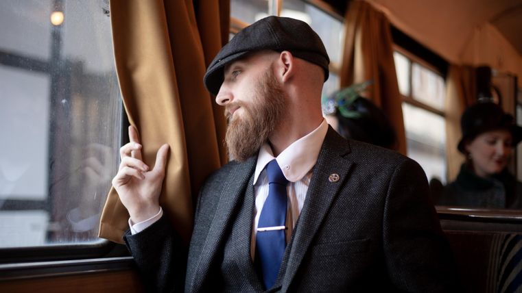 Ein gut gekleideter Mann mit einem Bart sitzt am Fenster eines Zugs. Er schaut nachdenklich nach draußen.