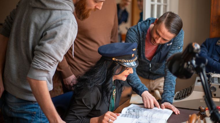 Gruppe von Menschen bei einem Krimispiel, eine Frau trägt eine Kapitänsmütze und betrachtet ein Buch.