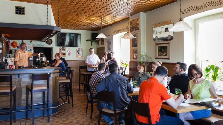 Gemütliches Restaurant mit Holztischen, Gästen und einem Barkeeper. Die Decke ist mit einem Muster verziert, Pflanzen stehen auf den Fensterbänken.