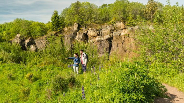 Zwei Wanderer stehen auf einem grünen Pfad vor der Felswand des Vulkangartens Steffeln.