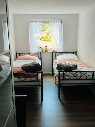 Een lichte slaapkamer met twee bedden en een raam dat veel daglicht binnenlaat. Op de vensterbank staan enkele bloemen in een vaas.