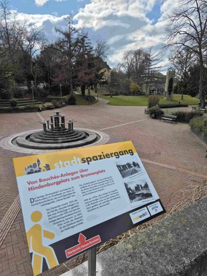 Ein Park mit einem Brunnen und einem Informationsschild über einen Stadtspaziergang.