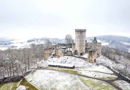 Eine massive Burg mit mehreren Türmen steht auf einer Felserhebung mitten in einer weitläufigen Wald- und Wiesenlandschaft im Winter. Die freien Wiesen sind leicht mit Schnee bedeckt.