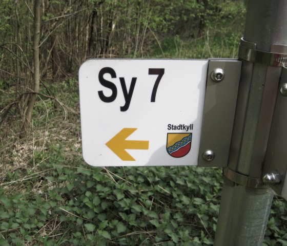 Wegwijzer in het bos met het opschrift 'Sy 7' en een gele pijl die naar links wijst. Daaronder staat het wapen van Stadtkyll., &copy; Touristik GmbH Gerolsteiner Land