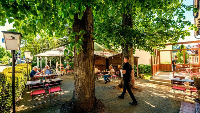 Un restaurant confortable en plein air avec des clients sous de grands arbres. Les tables sont couvertes de parasols et un serveur est en route.