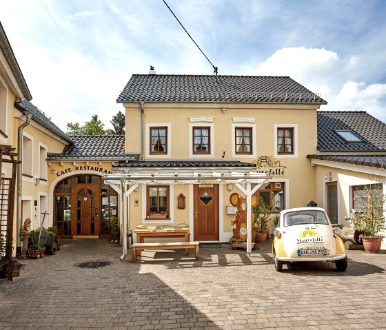 Yellow café called 'Mausefalle', decorated with plants. A small yellow car is parked in front of it. The sky is blue with a few clouds., © Christiane Weides
