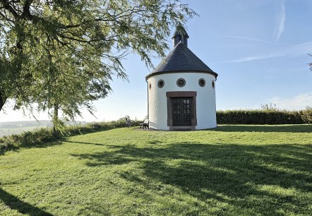 Round chapel with a black roof on a green meadow, surrounded by trees and blue sky. A bicycle leans against the chapel., &copy; Touristik GmbH Gerolsteiner Land