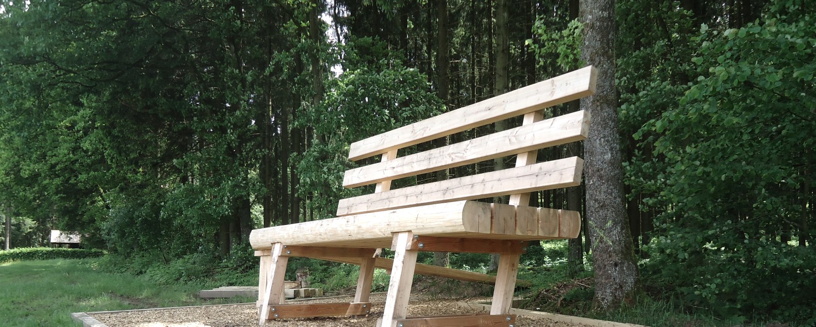 Eine &uuml;bergro&szlig;e Holzbank steht auf einem mit Holzschnitzeln bedeckten Platz im Wald, umgeben von B&auml;umen und Gras., &copy; Touristik GmbH Gerolsteiner Land