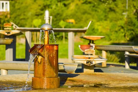 Ein Wasserspielplatz mit einem metallenen Brunnen, aus dem Wasser fließt. Im Hintergrund sind weitere Wasserspielelemente und grüne Vegetation zu sehen.