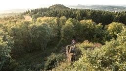 Twee mensen staan op een rots, omgeven door een dicht bos. Op de achtergrond zijn heuvels en een weids uitzicht over het landschap te zien., © Eifel Tourismus GmbH, D. Ketz