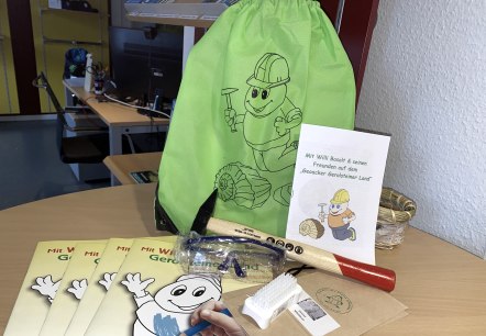 De uitrusting van de verkenner voor het geo-veld in Gerolstein ligt op een ronde houten tafel: een groene stoffen tas, verschillende kleurboeken, een hamer, een veiligheidsbril, een borstel en een informatieboekje.