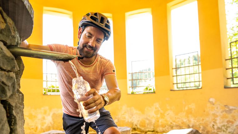 Ein Mann mit Fahrradhelm füllt an einem Brunnen in einem gelben Gebäude eine Wasserflasche auf