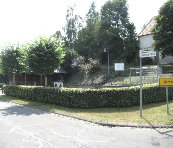 A village square with a barbecue hut, surrounded by trees and hedges. Road signs point to Ormont. In the background are trees and a building., © Touristik GmbH Gerolsteiner Land, Ute Klinkhammer