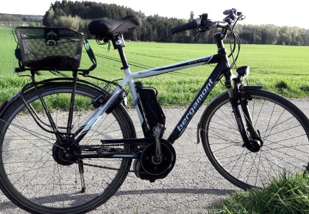 Ein Fahrrad geparkt am Rand eines asphaltierten Weges mit weiten Feldern und Wald im Hintergrund.