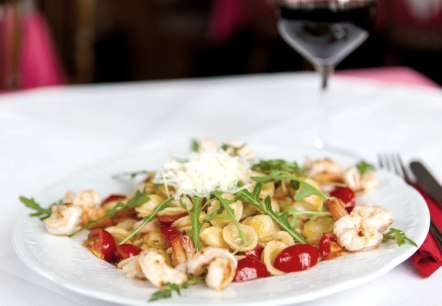 Auf einem Teller angerichtetes Nudelgericht mit Cherrytomaten, Garnelen, Rucola und Parmesan. Daneben auf einer Serviette liegendes Besteck und ein Glas Rotwein.
