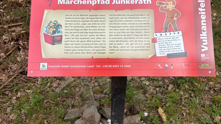 Eine Informationstafel des Märchenpfads Jünkerath im Wald, mit Text und einer Illustration eines Mannes mit Fernrohr.