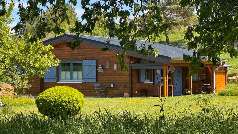 Ein gemütliches Holzhaus inmitten einer grünen Wiese. Umgeben von Bäumen und Sträuchern bietet es eine einladende Atmosphäre.