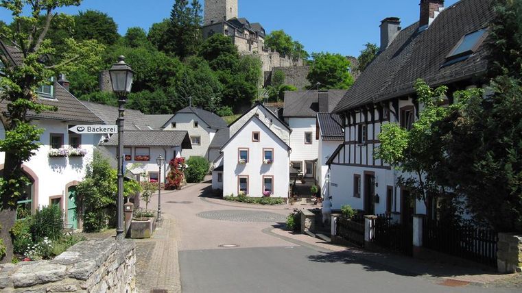 Eine malerische Dorfstraße mit weißen Häusern und grüner Vegetation. Im Hintergrund ist eine Burg zu sehen, die auf einem Hügel thront.