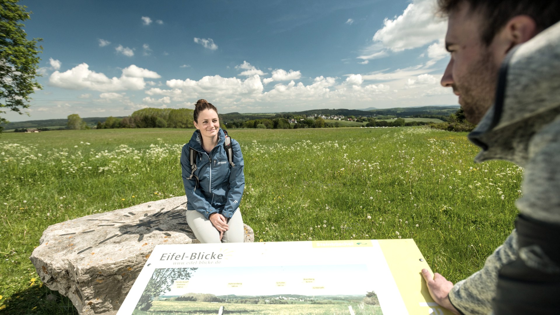 A woman is sitting on a stone with directional arrows on it. The stone stands on a large meadow, behind which lies a village. In the foreground is a man standing at an information board about the Eifel views and looking at the woman.