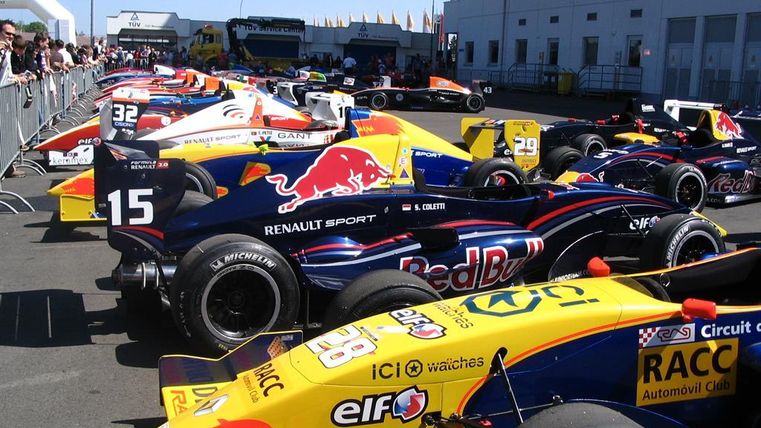 Eine Reihe von Rennwagen in bunten Farben, aufgestellt auf einer Rennstrecke. Im Hintergrund sind Zuschauer und Gebäude sichtbar.