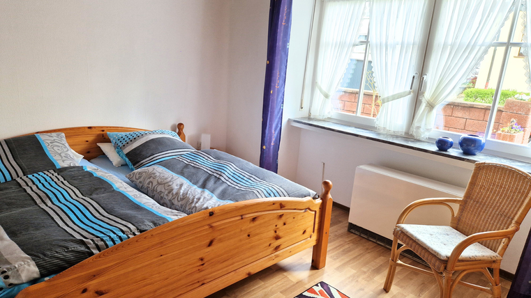 Une chambre lumineuse avec un lit en bois et des housses de couette colorées. Il y a une chaise et une fenêtre avec des rideaux, qui laisse entrer la lumière du jour.