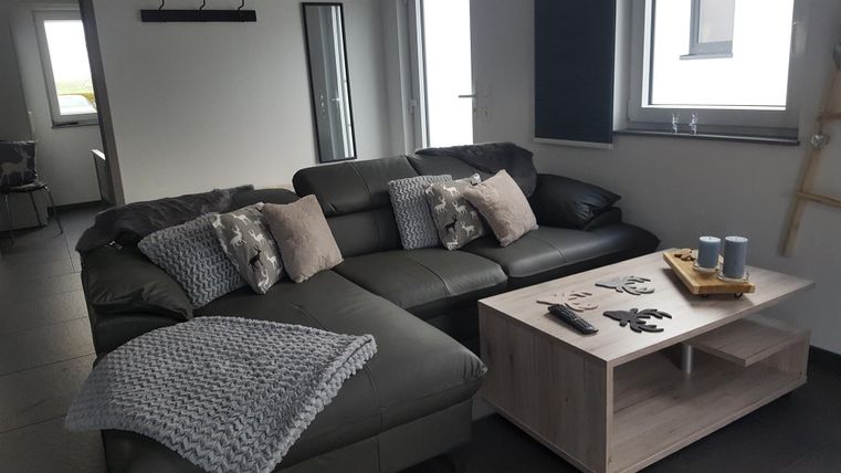 Ein modernes Wohnzimmer mit einem großen, grauen Sofa und dekorativen Kissen. Auf dem Tisch liegen eine Fernbedienung und ein Tablett mit Dingen.