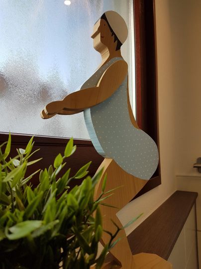 Eine stilisierte Figur steht an einem Fenster und schaut nach draußen. In der Nähe ist eine kleine Pflanze zu sehen.