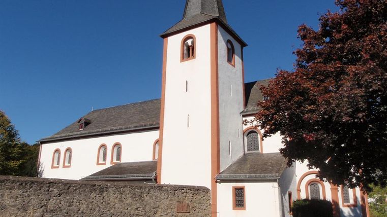 Eine alte Kirche mit einem hohen, spitzen Turm. Der sonnige Himmel und die umgebenden Bäume verleihen der Szene einen ruhigen Charakter.