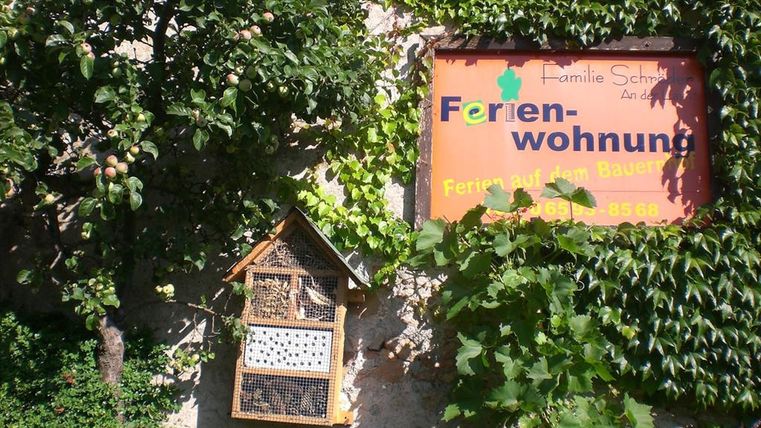 Ein gemütliches Ferienwohnungen-Schild umgeben von grünen Pflanzen. Neben dem Schild steht ein Insektenhotel.